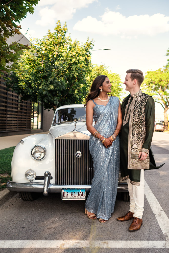 Bride and groom in traditional Indian attire stands in front of a vintage white car on a sunny street during their Sangeet at Ovation Chicago