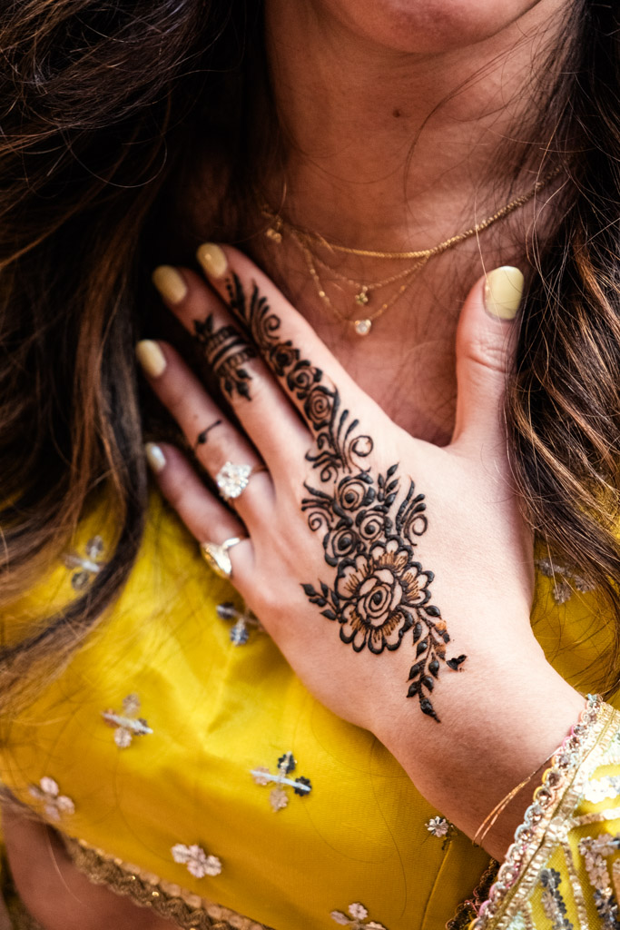 Hand with floral henna design rests on chest; person wears yellow outfit and gold jewelry