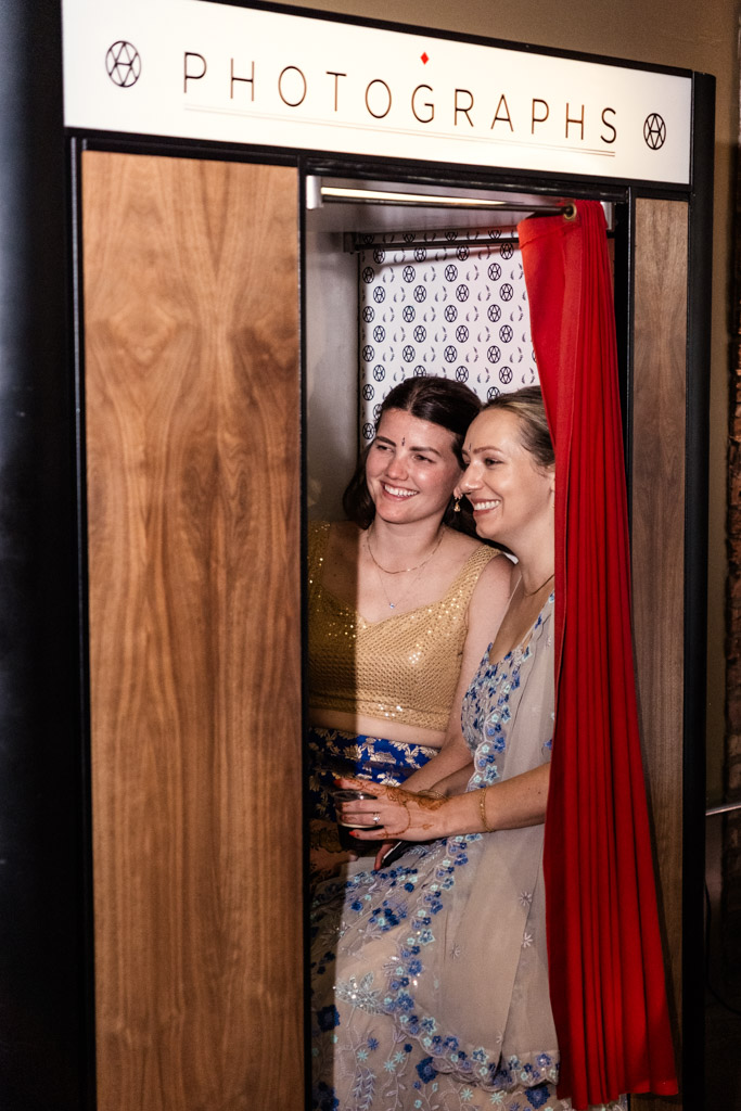 Two women in colorful dresses smile together inside a vintage photo booth with a red curtain during Sangeet at Ovation Chicago