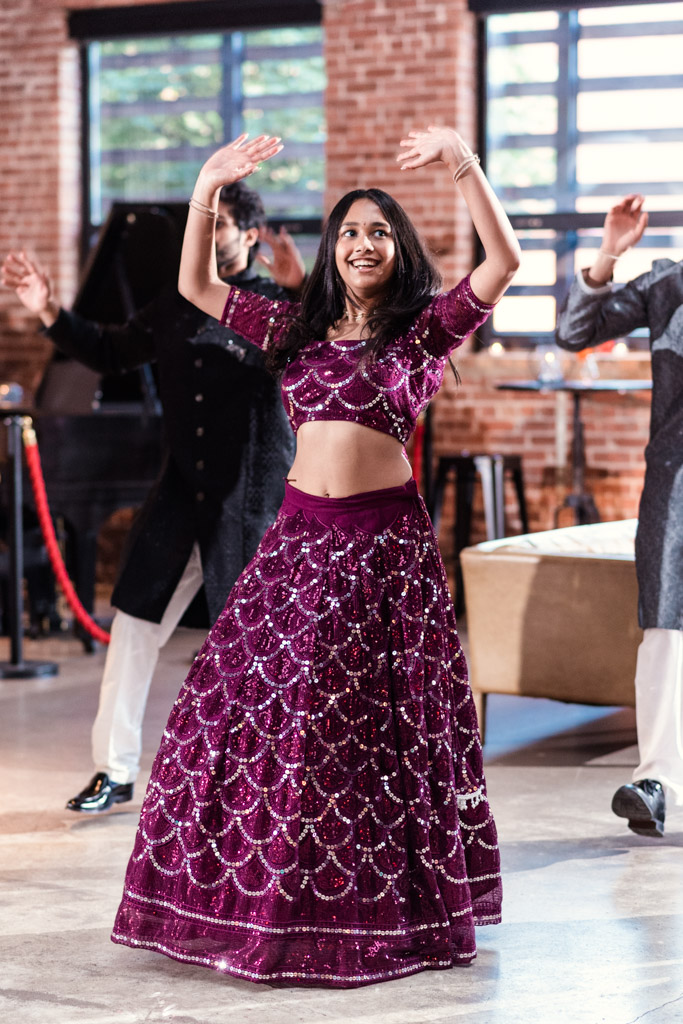 Woman in a purple, sequined lehenga dances indoors at Chicago Sangeet, arms raised, smiling, with others in the background