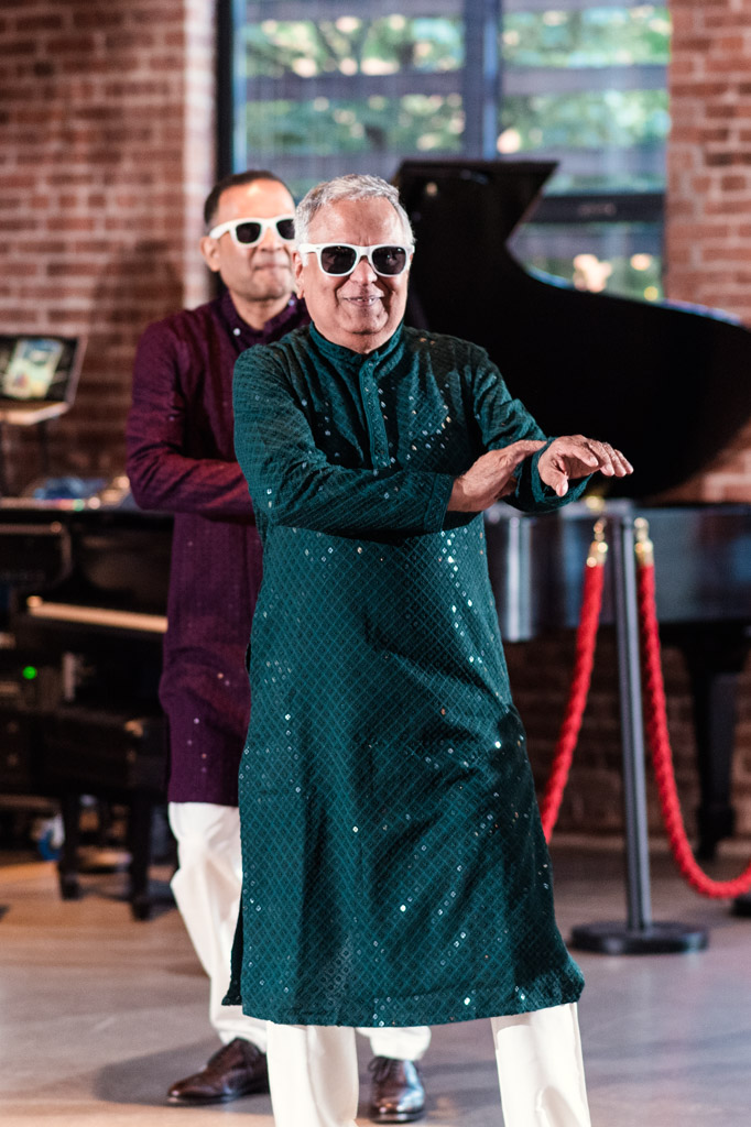 Two men in festive attire and sunglasses dance indoors during a Sangeet at Ovation Chicago, with a piano and red rope in the background