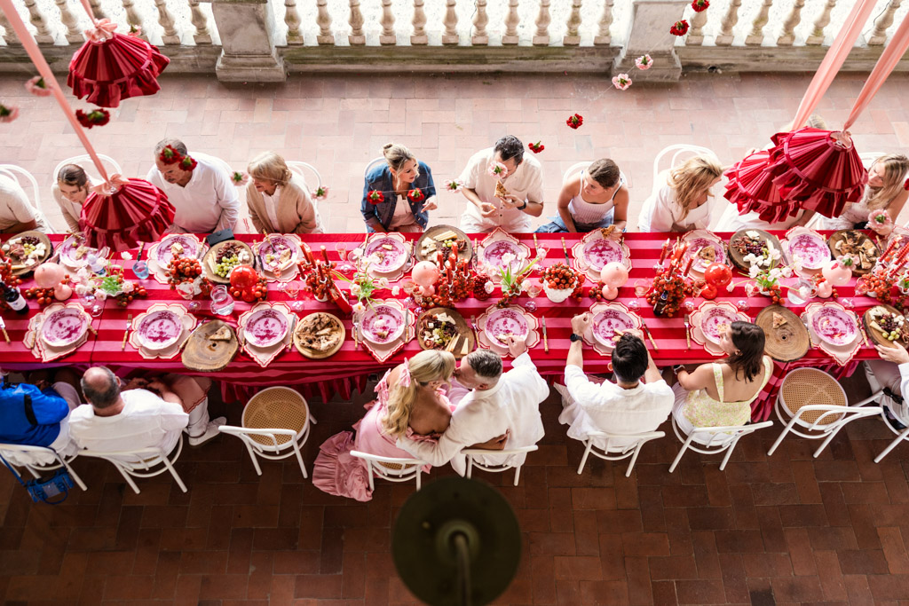 Photo from above of guests gathered at one long dinner table in Italian Villa for a destination wedding welcome party with pizza and pink and red decor