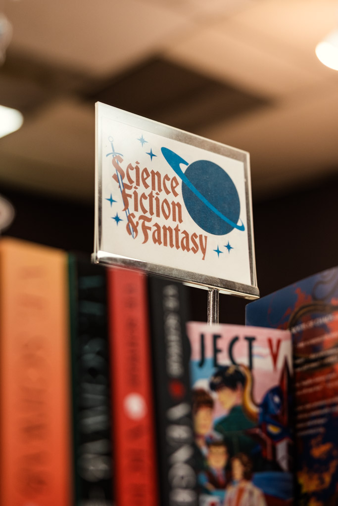 A sign reading “Science Fiction & Fantasy” is displayed above a row of colorful books on a shelf, inviting bookstore engagement from curious readers exploring new worlds.