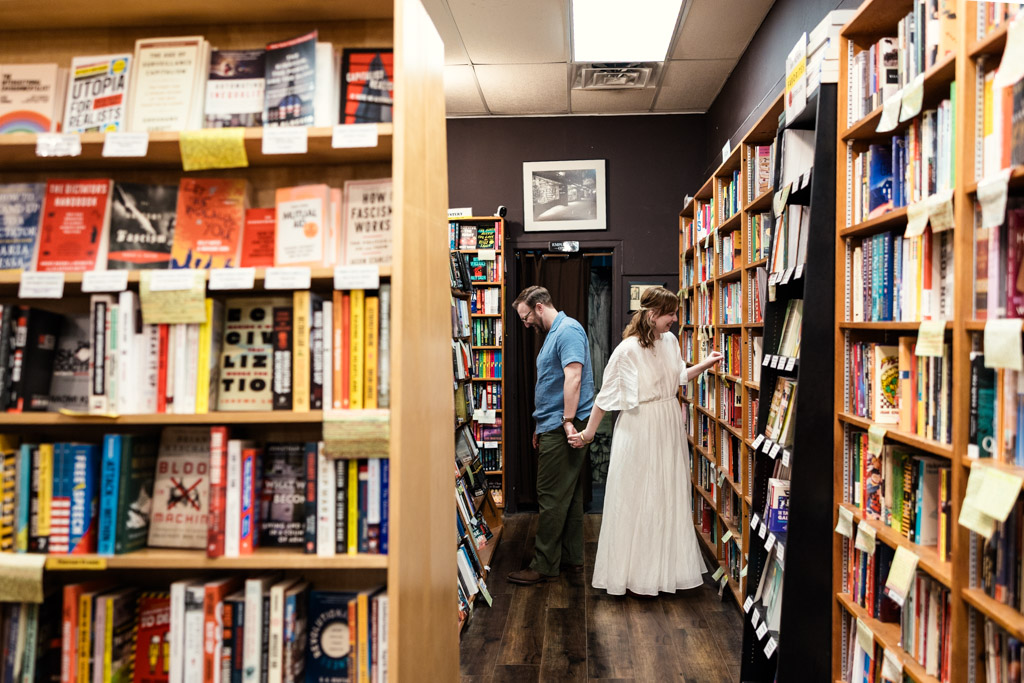 A couple stands back to back in a bookstore aisle, browsing shelves filled with colorful books during their cozy bookstore engagement.
