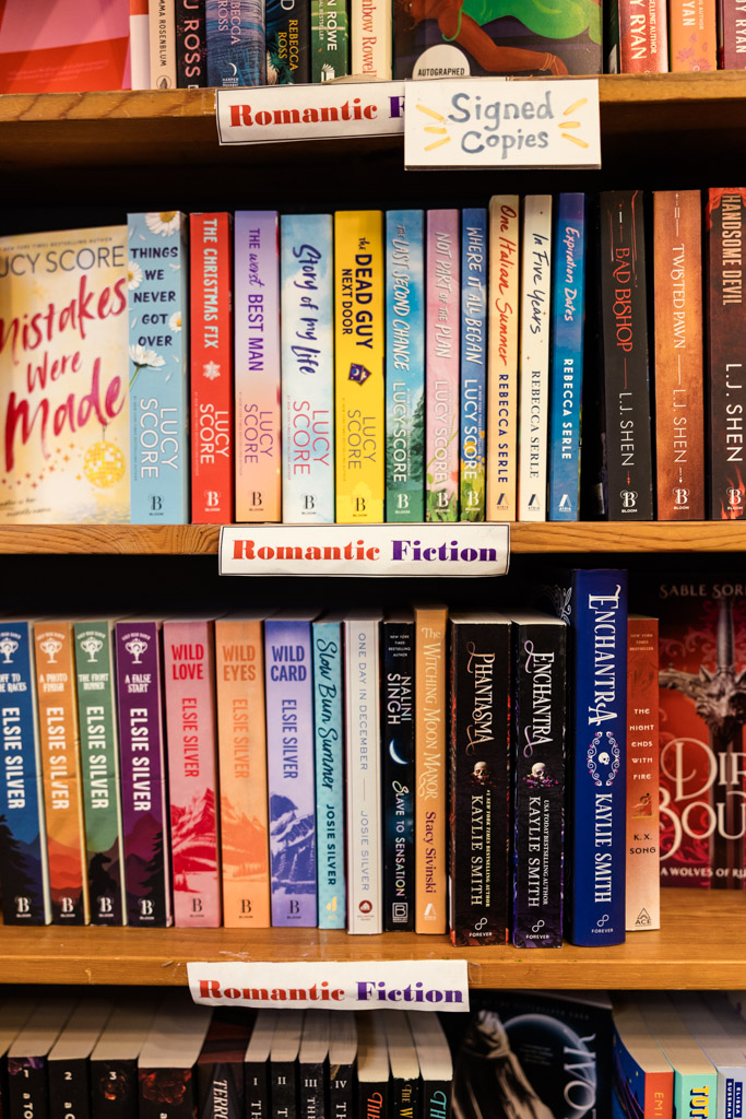Bookshelves labeled “Romantic Fiction” with colorful books and a “Signed Copies” sign on the top shelf invite bookstore engagement for every hopeless romantic wandering the aisles.