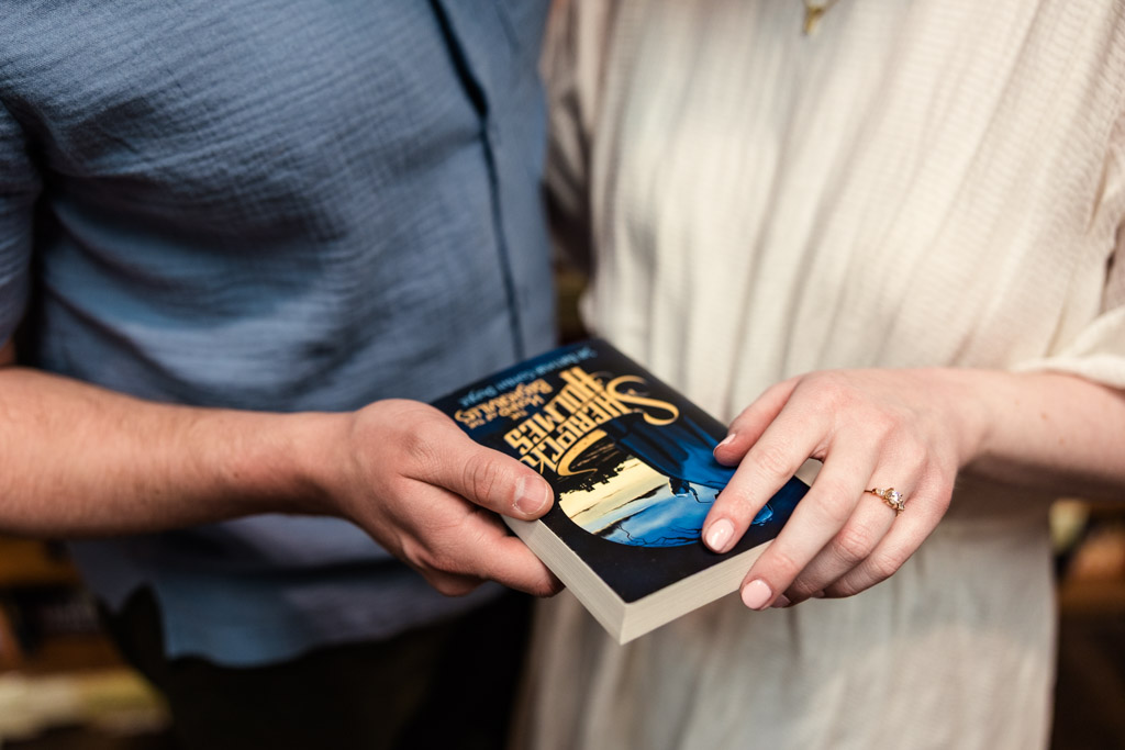 Two people in light clothing hold a book together, their hands and the book cover in clear focus, capturing a moment of bookstore engagement.