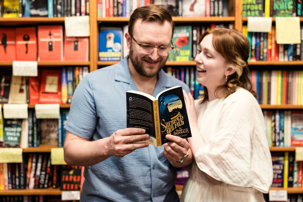 A man and woman smile while reading a Sherlock Holmes book together during their bookstore engagement.