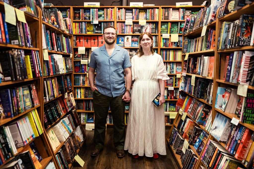 Two people stand holding hands in a bookstore aisle, surrounded by colorful shelves of books, sharing a quiet moment during their bookstore engagement.