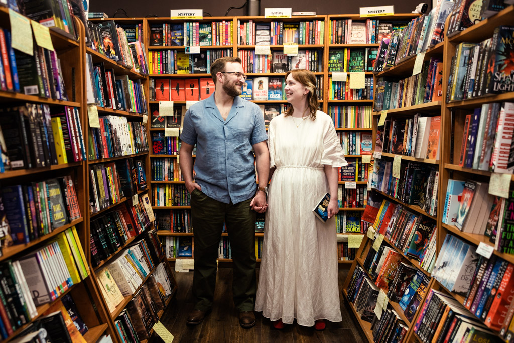 A couple enjoying a bookstore engagement holds hands and smiles at each other in a colorful bookstore aisle filled with books.