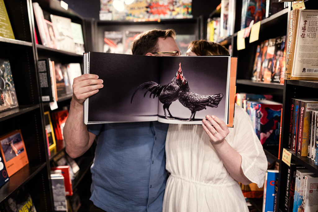 Two people hide their faces behind an open book showing a large photo of a black rooster in a charming bookstore, capturing a playful moment of bookstore engagement.