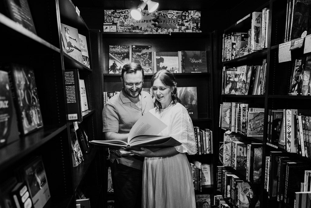 A couple stands close together, smiling while reading a large book in a bookstore aisle, capturing the warmth of a charming bookstore engagement.
