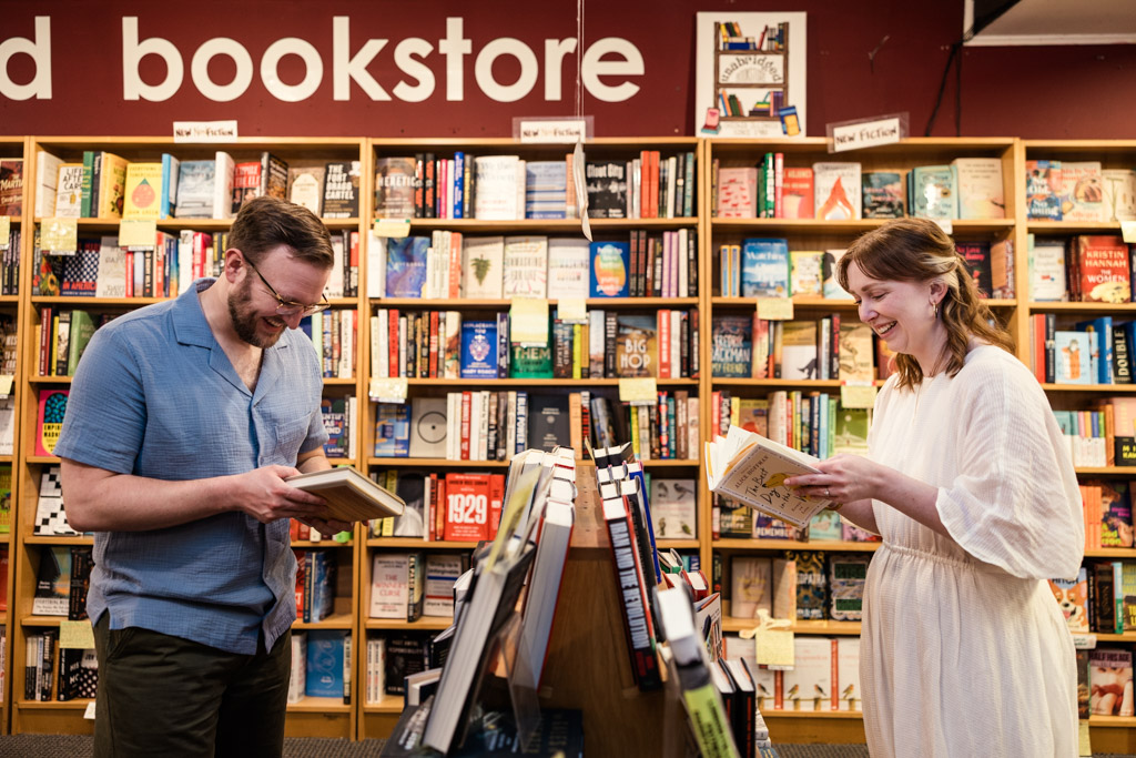 Two people smiling and reading books in a bookstore, enjoying a moment of bookstore engagement as they’re surrounded by shelves filled with colorful books.
