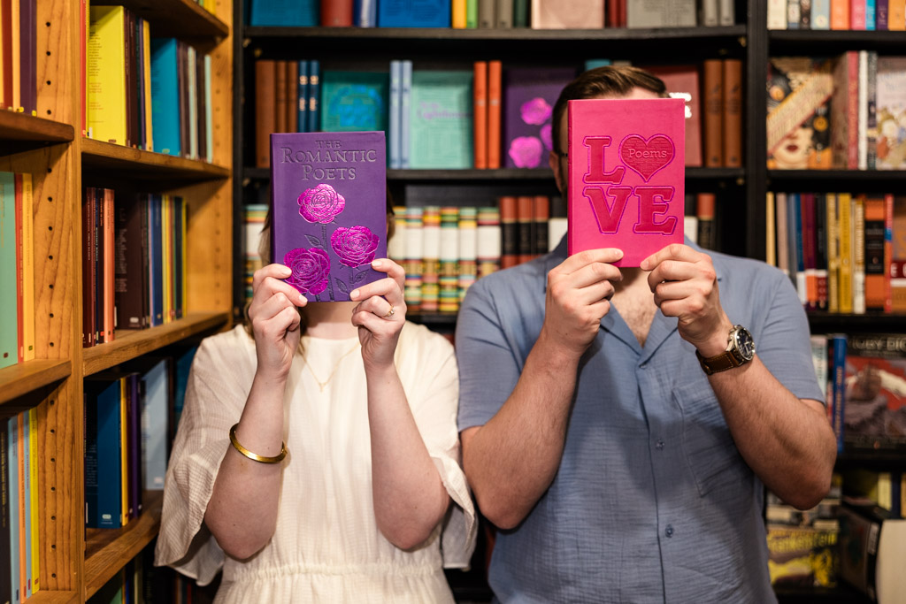 Two people holding poetry books over their faces in a colorful bookstore, standing close together, capture the whimsical spirit of a bookstore engagement.