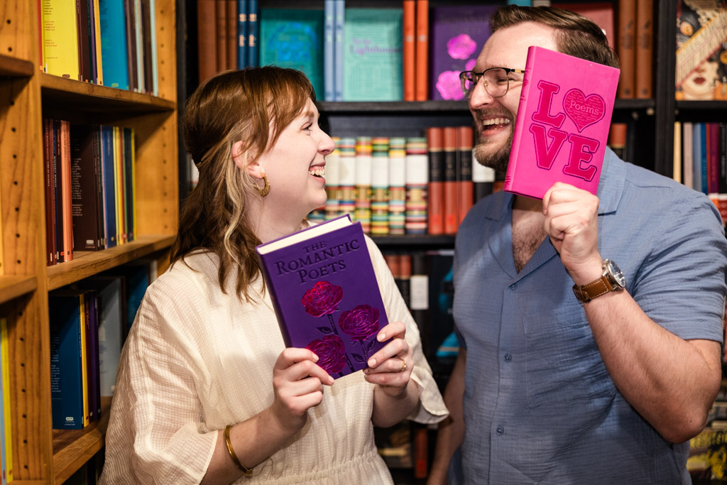 Two people smile at each other in a library, each holding a bright pink book with romantic themes, capturing a charming bookstore engagement vibe.
