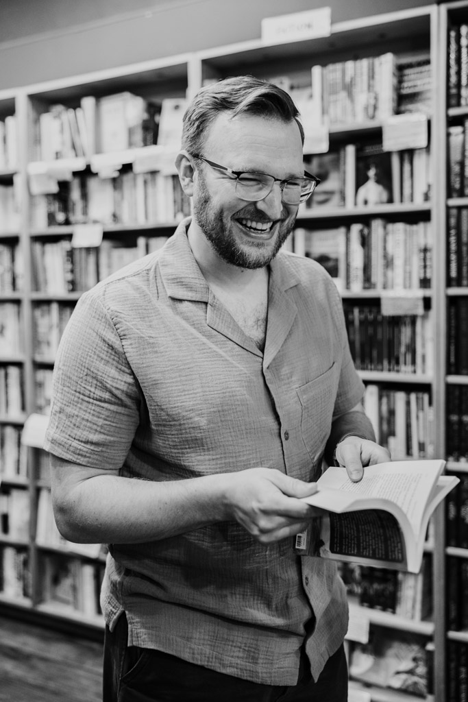 Man smiling while holding an open book in a bookstore, enjoying genuine bookstore engagement as he stands surrounded by shelves filled with books.