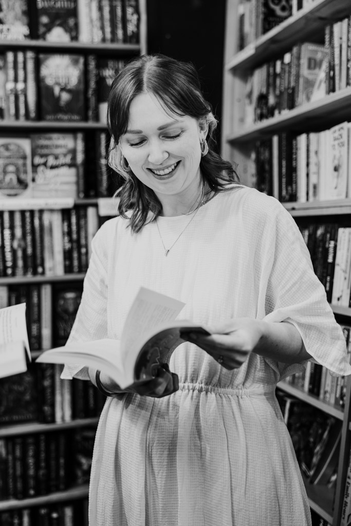 A woman in a light dress smiles while reading a book during her bookstore engagement, surrounded by shelves in a cozy library or bookstore.
