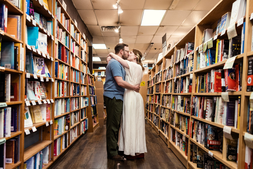A couple shares an intimate embrace and kiss in the aisle of a bookstore lined with shelves of books, capturing a heartfelt bookstore engagement.