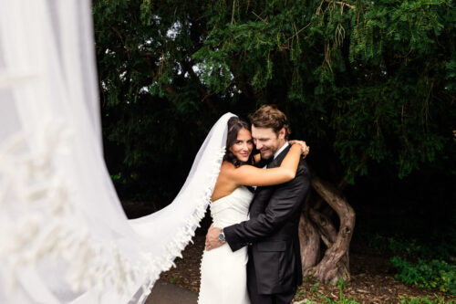 Bride and groom embrace outdoors at Cafe Brauer, her veil flowing with green trees in the background.