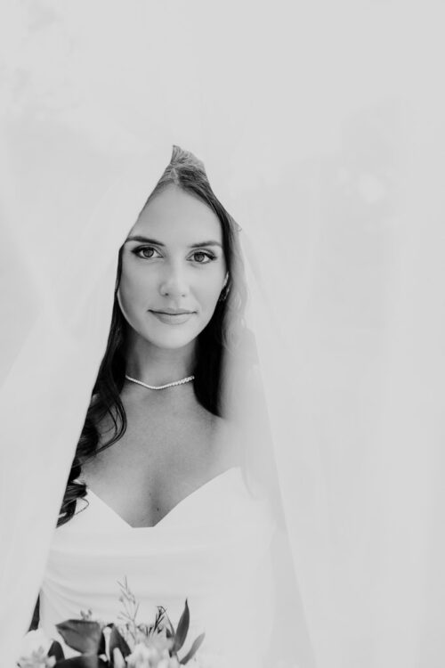 A bride in a strapless dress poses under a sheer veil at Cafe Brauer, holding a bouquet, in black and white.