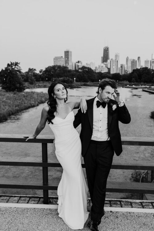 A bride and groom pose on a bridge near Cafe Brauer with a city skyline in the background, looking relaxed.