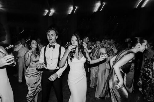 A lively group dressed formally, dancing and holding drinks at a Cafe Brauer party or wedding reception.