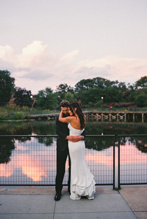 Bride and groom embrace by a lake at sunset near Cafe Brauer, with trees and clouds reflected in the water.