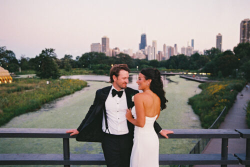 A bride and groom stand on a bridge near Cafe Brauer, smiling with the city skyline in the background.