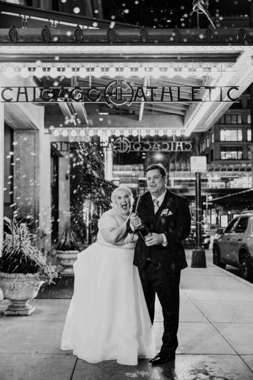 A couple in wedding attire laughs popping champagne outside Chicago Athletic Association at night.