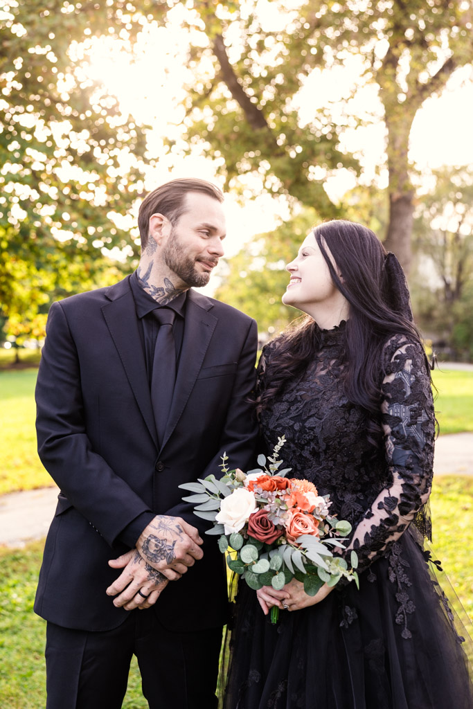 A couple in formal black wedding attire looking at each other while holding a bouquet.
