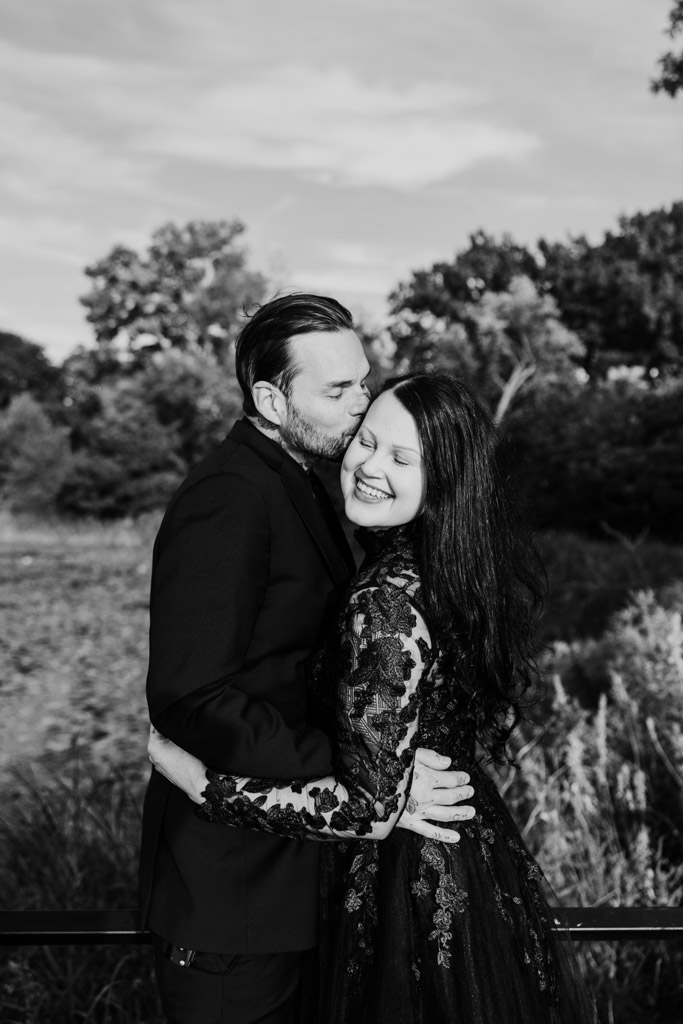 A man in a suit kisses a smiling woman in a black lace dress outdoors.