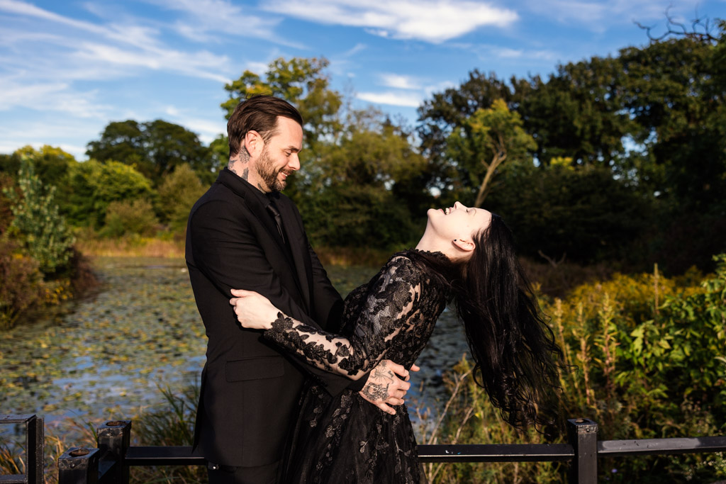 A man in a black suit holds a woman in a black lace dress who is leaning back and laughing outdoors.