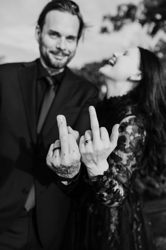A bride and groom laughing while holding up their middle fingers to show off their wedding rings.
