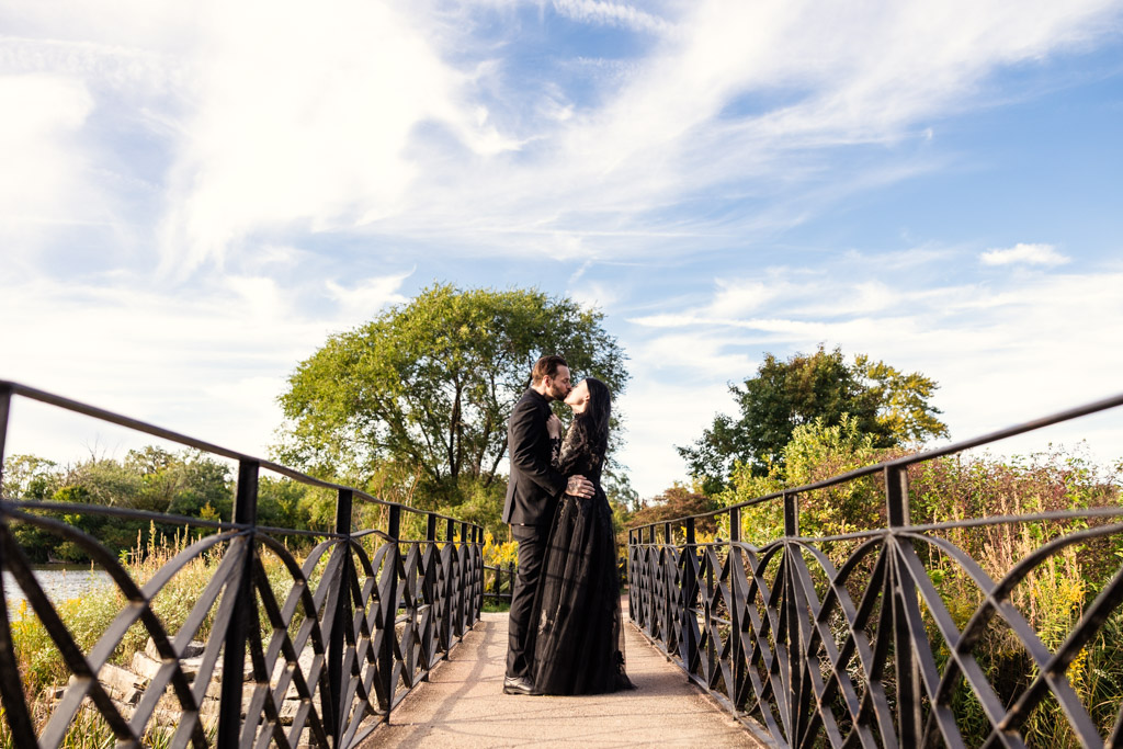 A couple in black wedding attire kissing on a bridge.