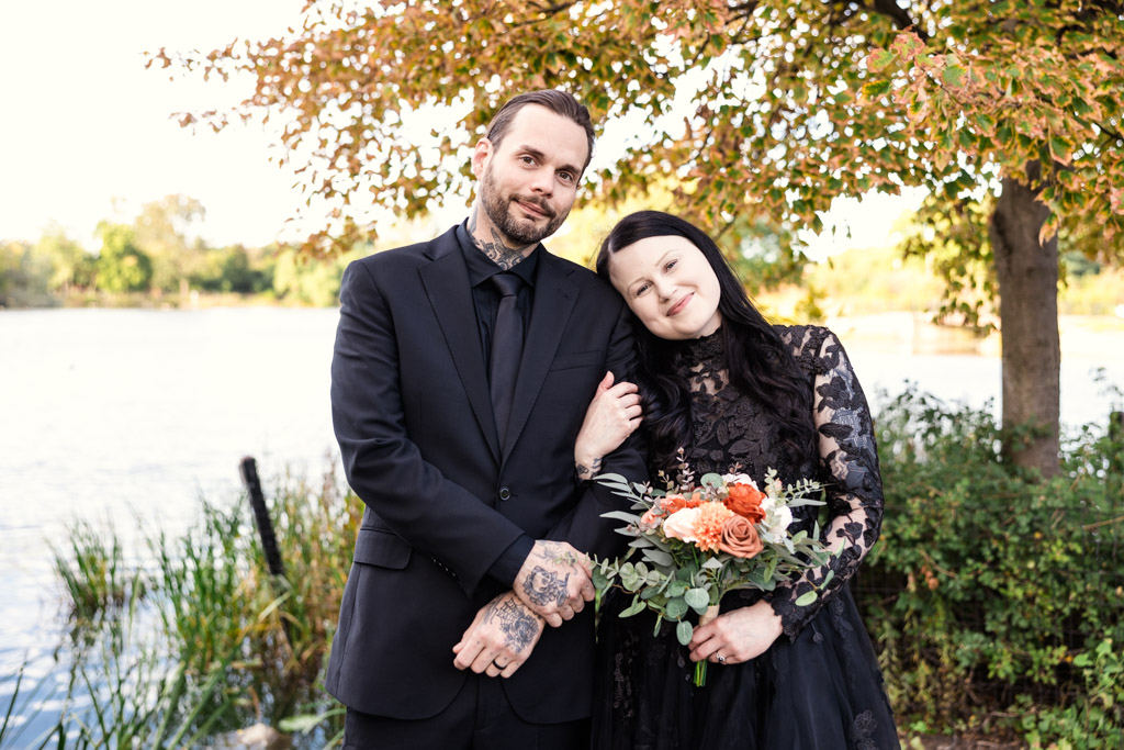 A couple in formal black attire pose together outdoors near a lake, with the woman holding a bouquet.