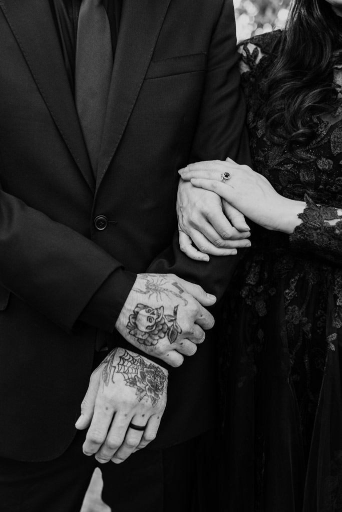 A person in a black suit with tattooed hands holding the arm of someone wearing a black lace dress.