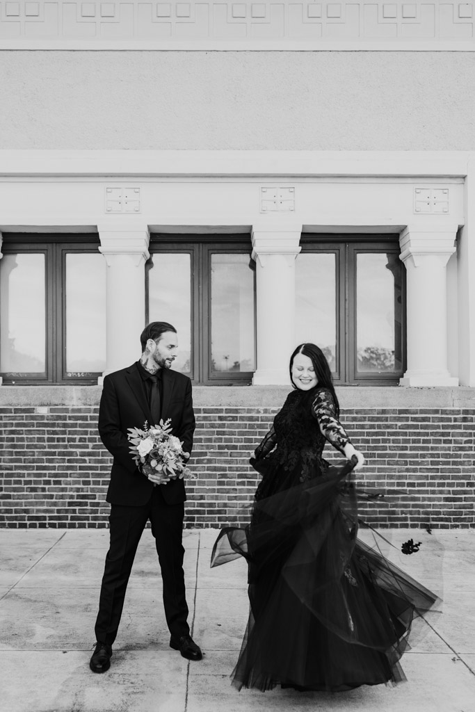A person in a black suit holds a bouquet while a person in a black gown spins in front of a brick building.