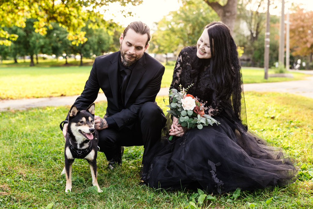 A couple in black wedding attire kneeling in a park with their Shiba Inu dog.