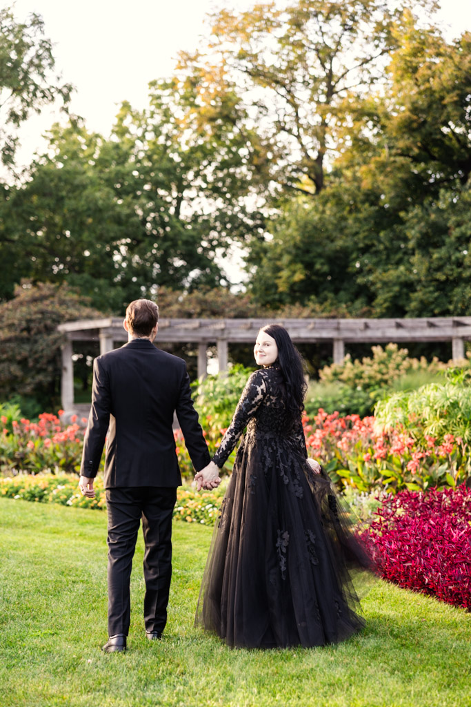 A couple in formal black attire holding hands while walking through a garden.