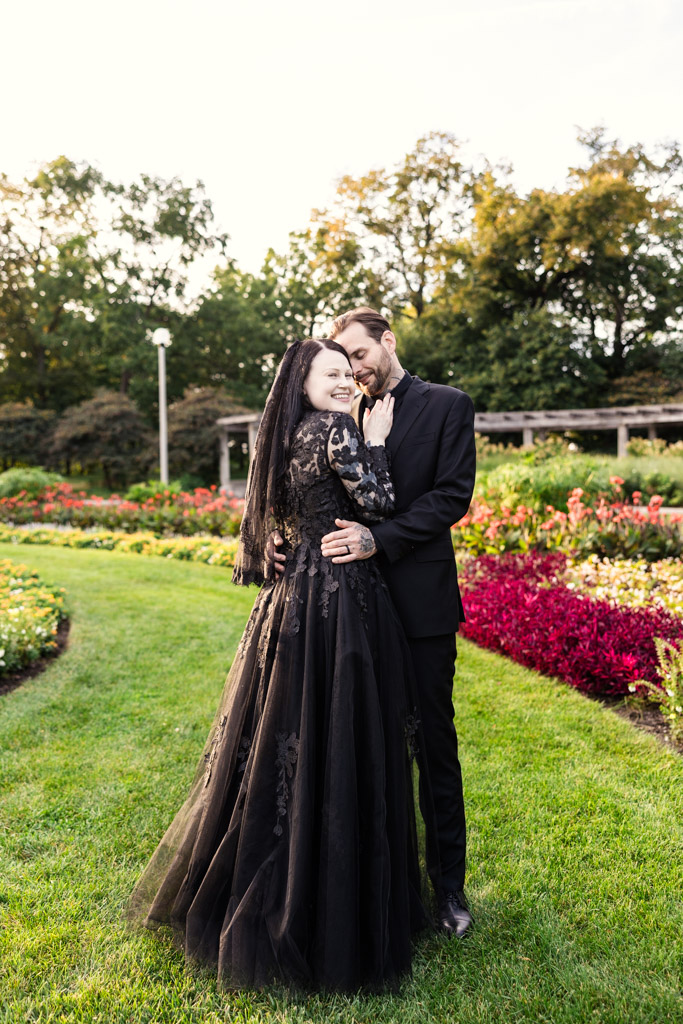 A bride in a black lace gown and a groom in a black suit embrace in a garden.