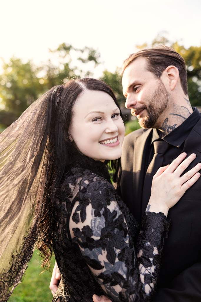 A bride in a black lace dress and veil smiling at the camera next to her partner in a black suit.