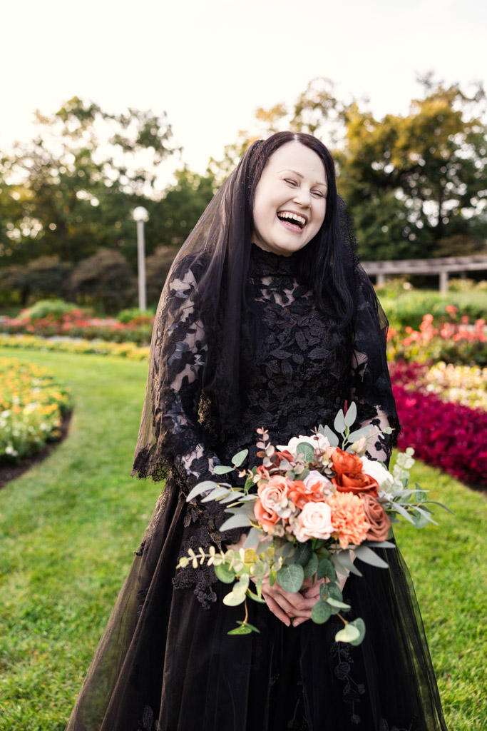 A laughing bride in a black wedding dress and veil holds a bouquet of flowers in a garden.