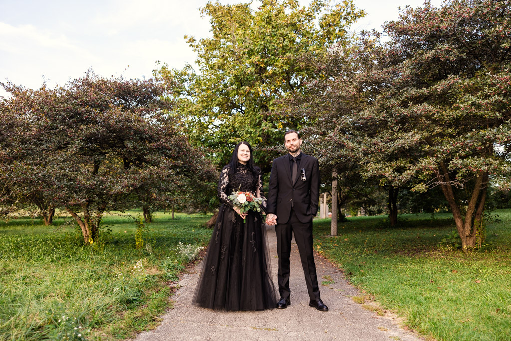 A bride in a black wedding dress and a groom in a black suit standing together on a park path.