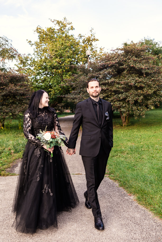 A couple in black wedding attire walking hand-in-hand through a park.