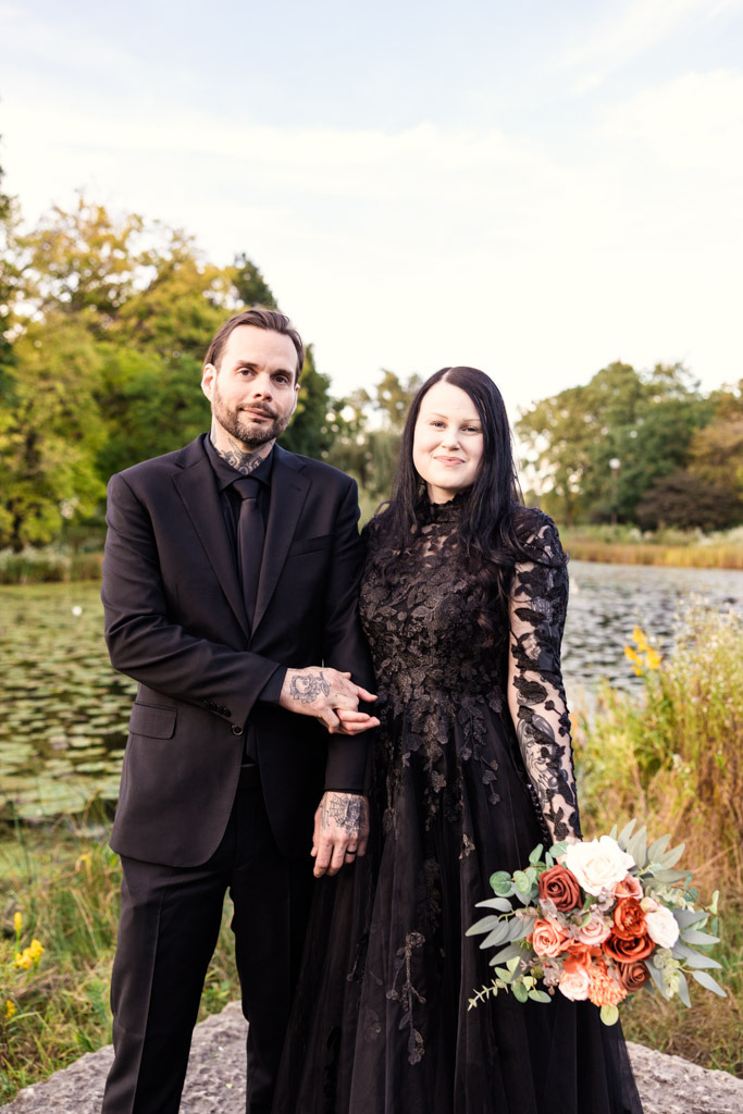 A bride and groom in black wedding attire holding hands by a pond.