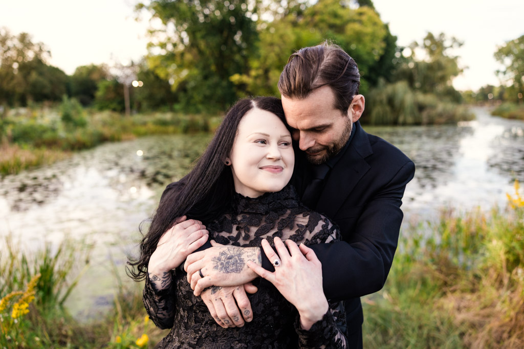 A man in a black suit embraces a woman in a black wedding dress by a pond.