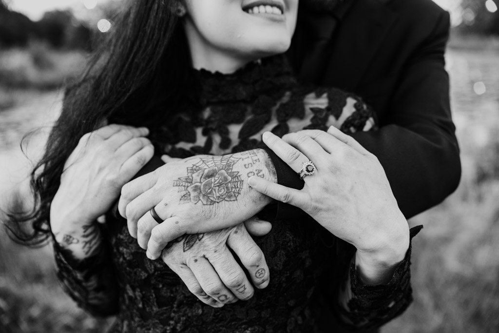 A person in a black wedding dress is embraced from behind, showcasing their wedding rings and hand tattoos.