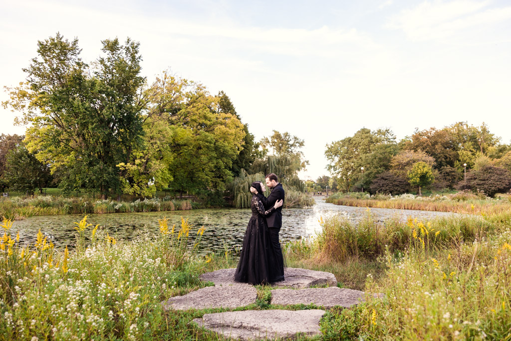 A couple in formal attire embracing on stone steps beside a pond.