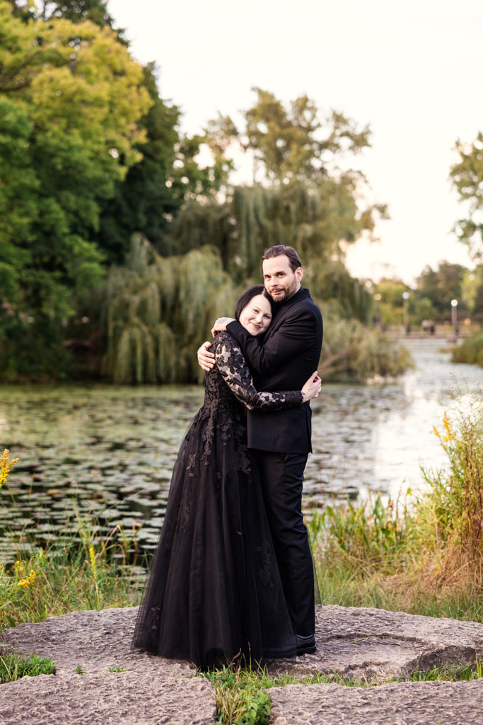 A couple in formal black attire embracing on a rock by a pond.