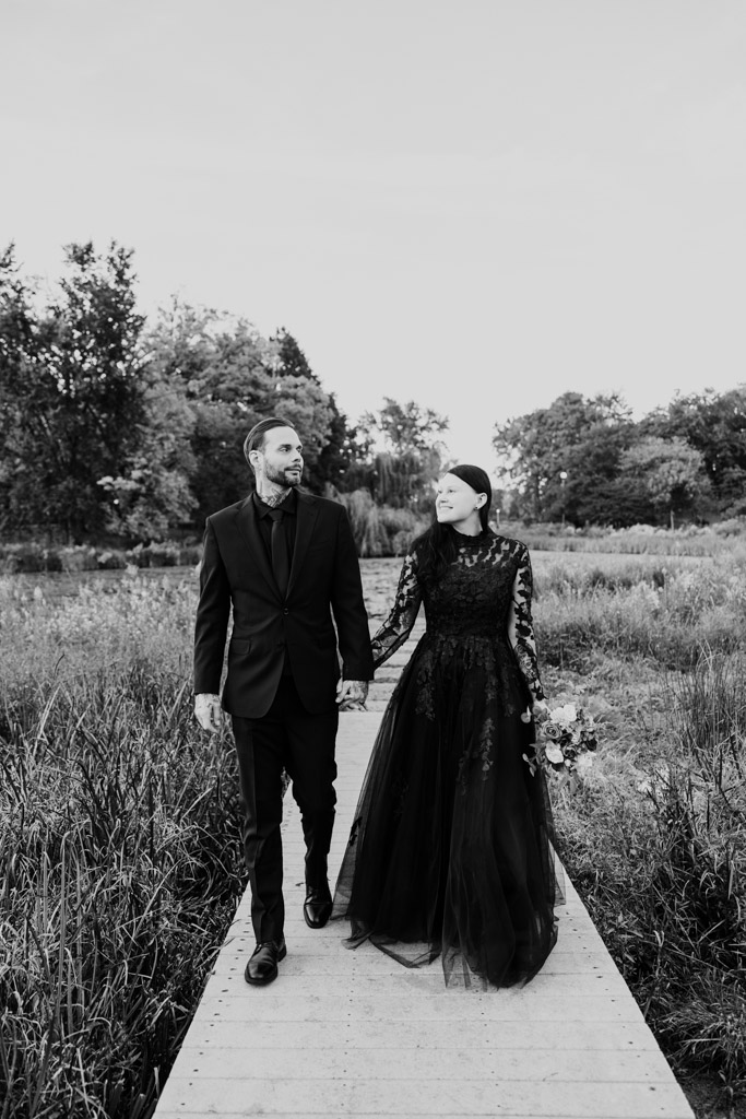 A bride and groom in black wedding attire walking hand-in-hand along a wooden boardwalk in a meadow.