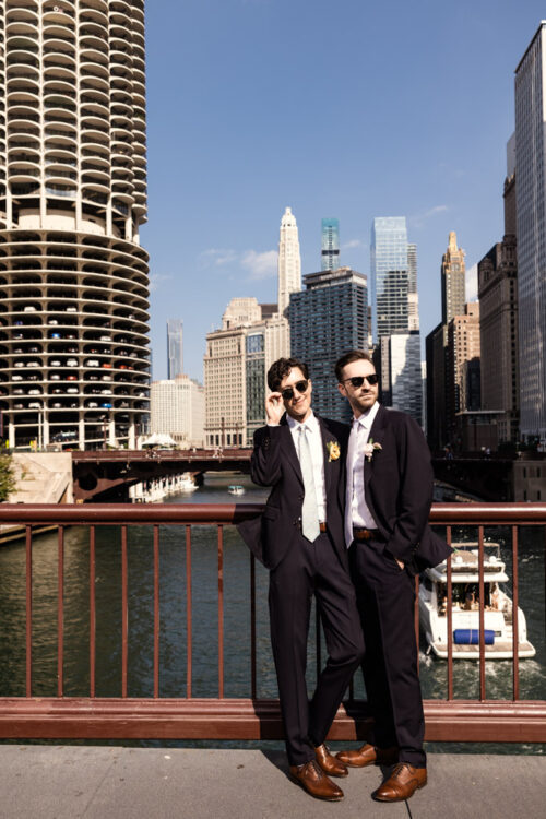 Two grooms in dark suits and sunglasses standing on a Chicago bridge with the skyline in the background.